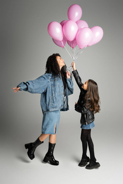 Vista lateral de la elegante madre y su hija sosteniendo globos rosados sobre fondo gris  - Foto, imagen