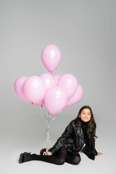 Χαμογελώντας preteen παιδί σε δερμάτινο μπουφάν κοιτάζοντας κάμερα κοντά σε ροζ μπαλόνια σε γκρι φόντο  - Φωτογραφία, εικόνα