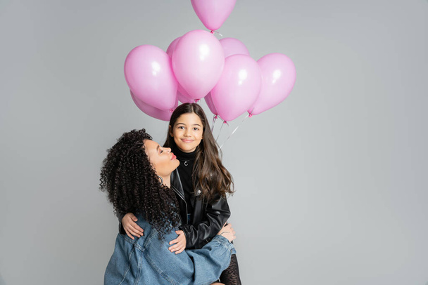 Mulher encaracolada abraçando a filha sorridente perto de balões cor-de-rosa isolados em cinza  - Foto, Imagem