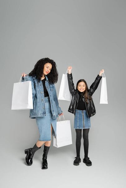 Full length of stylish mother and daughter holding shopping bags on grey background  - Valokuva, kuva