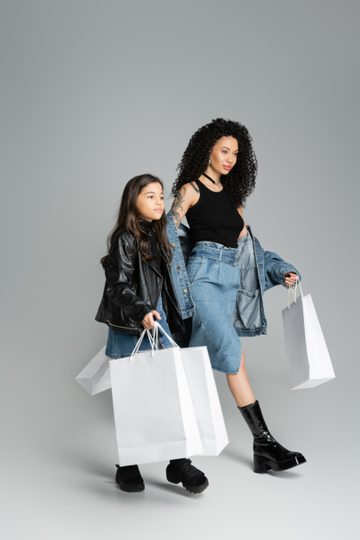 Trendy mother and child holding shopping bags and walking on grey background  - Foto, imagen