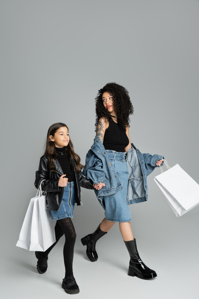 Trendy μαμά και η κόρη κρατώντας τα χέρια και τσάντες ψώνια σε γκρι φόντο  - Φωτογραφία, εικόνα