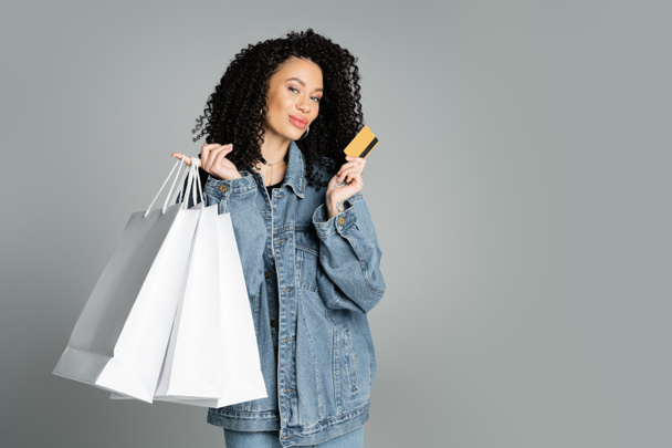 Модна жінка в джинсовій куртці тримає кредитну картку і сумки для покупок ізольовані на сірому
  - Фото, зображення