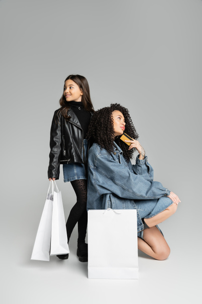 Pozitív tizenéves lány gazdaság bevásárló táskák közelében anya hitelkártya szürke háttér  - Fotó, kép