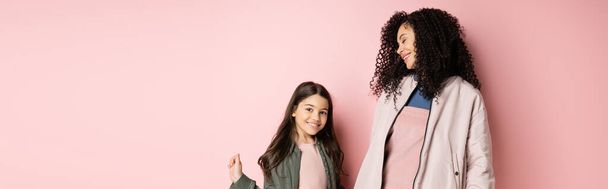 Curly woman smiling near stylish daughter on pink background, banner  - 写真・画像