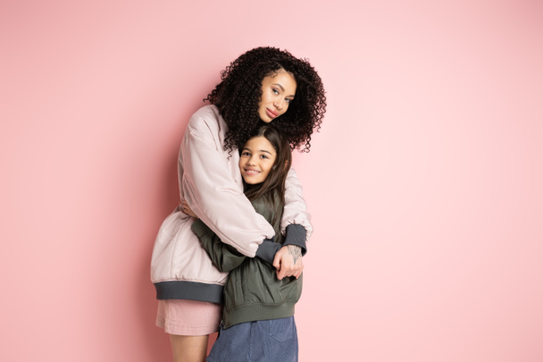 Lockige Frau in Bomberjacke umarmt Tochter auf rosa Hintergrund  - Foto, Bild