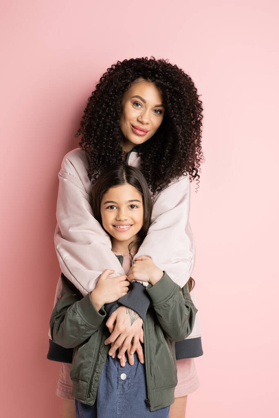 Curly woman hugging preteen girl on pink background  - 写真・画像
