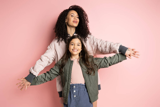 Curly mother holding hands of cheerful daughter on pink background  - Foto, imagen