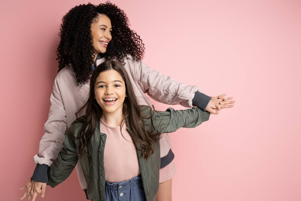 Positive curly woman holding hand of preteen daughter on pink background  - 写真・画像
