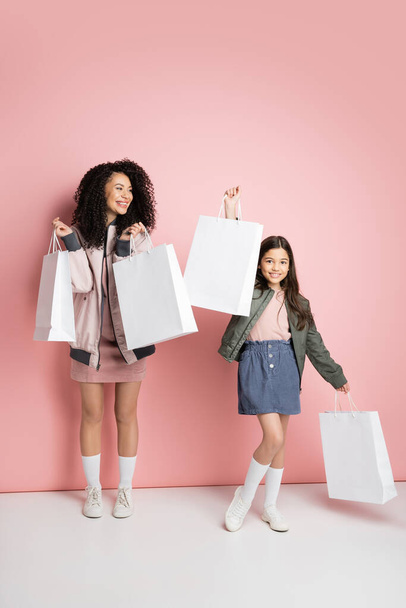 Cheerful mother and daughter holding shopping bags on pink background  - Фото, изображение