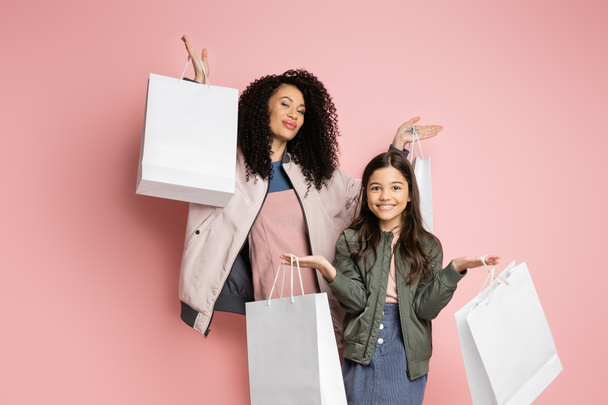Fashionable mom and daughter holding shopping bags and looking at camera on pink background  - Photo, Image