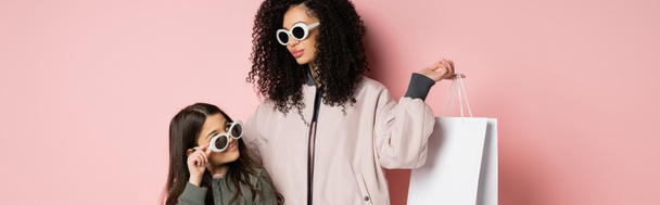 Madre de moda en gafas de sol con bolsa de compras cerca de la hija sobre fondo rosa, pancarta  - Foto, Imagen