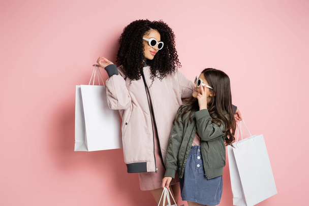 Trendy woman and kid in sunglasses holding shopping bags on pink background  - Photo, Image