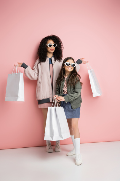 Full length of trendy mother and daughter in sunglasses holding purchases on pink background  - Foto, immagini