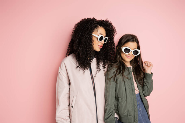 Donna e ragazza alla moda in occhiali da sole in piedi su sfondo rosa  - Foto, immagini
