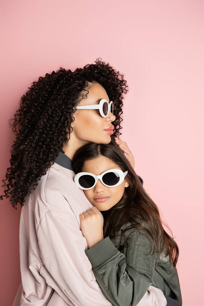 Trendy woman in sunglasses hugging preteen kid on pink background  - Photo, image