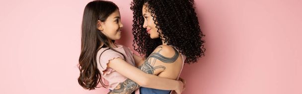 Vista lateral del niño sonriente abrazando a la mamá tatuada sobre fondo rosa, pancarta  - Foto, imagen