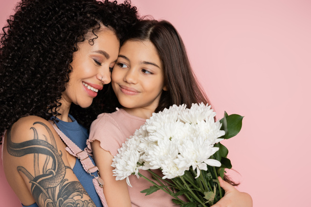 Sonriente mamá tatuada abrazando al niño con flores blancas aisladas en rosa  - Foto, imagen