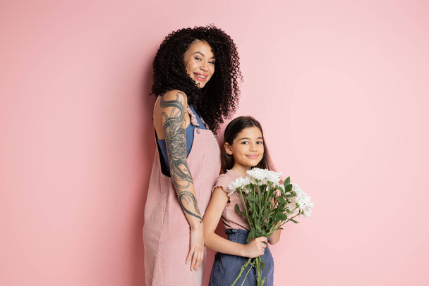 Fashionable woman looking at camera near daughter with flowers on pink background  - Photo, Image