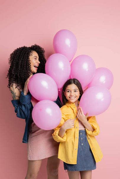 Positive mother and stylish kid standing near festive balloons on pink background  - Foto, Imagen
