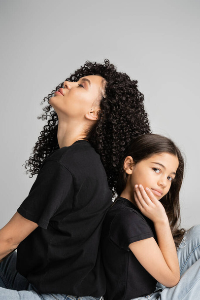Curly mother and daughter in black t-shirts sitting back to back isolated on grey - Φωτογραφία, εικόνα