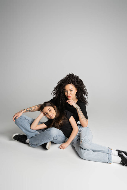Tattooed woman and daughter in jeans and t-shirts smiling at camera on grey background  - Foto, Bild