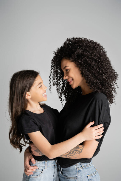 Side view of positive woman and daughter in black t-shirts hugging isolated on grey  - Fotó, kép