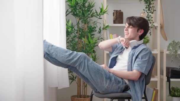 Adolescente chico enviando mensaje de voz en reloj inteligente mientras se relaja sentado en la silla al lado de la ventana - Metraje, vídeo