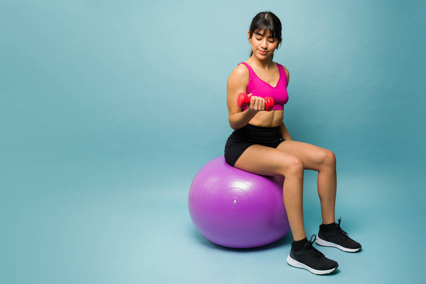 Mujer fuerte en forma sentada en una bola de estabilidad y levantando pesas de mancuerna mientras entrena y hace ejercicio al lado del espacio de copia - Foto, imagen