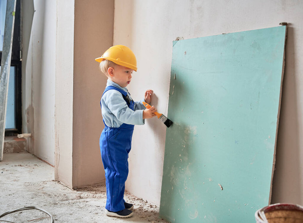 Little boy construction worker painting wall with brush in apartment under renovation. Child wearing safety helmet and work overalls while playing at home. - Photo, Image