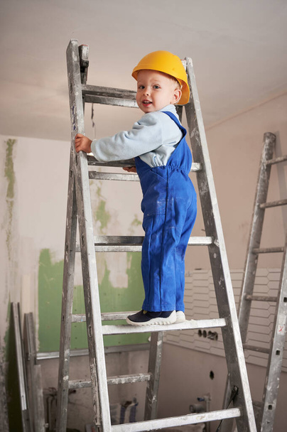 Child construction worker on the top of ladder while working on home renovation. Kid in safety helmet and work overalls walking up staircase in apartment under renovation. - Foto, Imagen