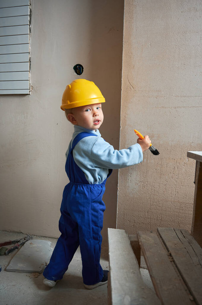 Boy construction worker painting wall with brush in apartment under renovation. Child wearing safety helmet and work overalls while playing at home. - Fotoğraf, Görsel