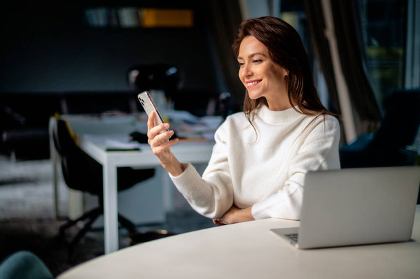 Middle aged business woman text messaging and using laptop while sitting at desk at the office. Executive businesswoman wearing sweater.  - 写真・画像