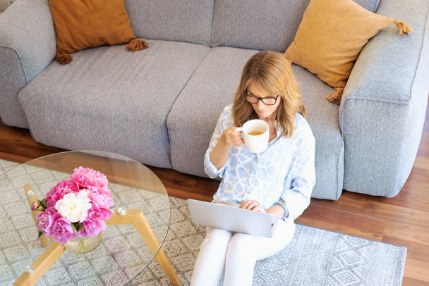 Middle aged woman sitting at home and using laptop for work. Blond haired businesswoman wearing casual clothes and drinking tea while working at home. Home office.  - Foto, imagen