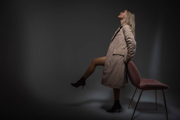 junges, schönes, emotionales Mädchen in grauem Regenmantel steht neben einem Stuhl, erhebt ein Bein, lehnt auf weißem Hintergrund, ein Lichtstrahl - Foto, Bild