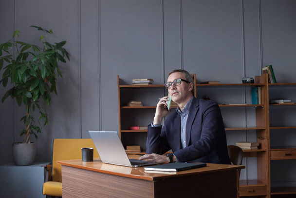 Hombre de negocios de edad avanzada de pelo gris en gafas que trabajan en el ordenador portátil mientras habla en el teléfono móvil. - Foto, imagen