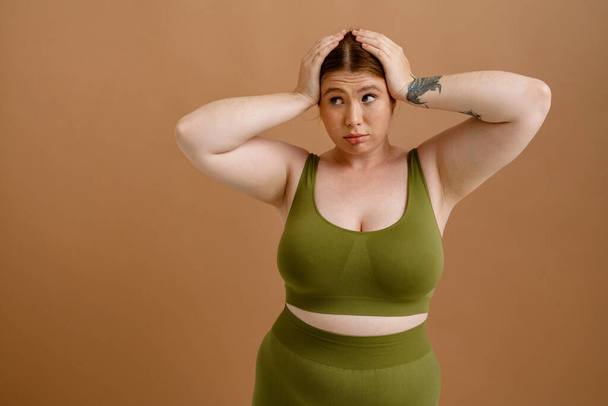 Young beautiful tattoed plus size girl in training suit holding her head and looking aside , while standing over isolated brown background - Photo, image