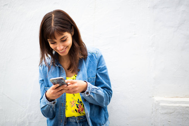 Portrait of smiling young woman leaning against white wall and using cellphone - Φωτογραφία, εικόνα