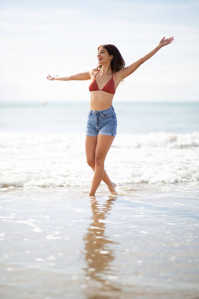 Full body portrait of excited young woman walking on the beach with arms outstretched - Foto, afbeelding