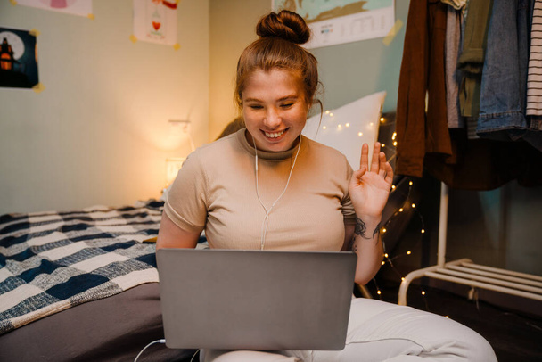 Πορτρέτο του νεαρού όμορφο χαμογελαστό κορίτσι σε ακουστικά που κατέχουν συνέντευξη βίντεο με φορητό υπολογιστή και χαιρετισμό, ενώ κάθεται στο άνετο δωμάτιο στο σπίτι - Φωτογραφία, εικόνα
