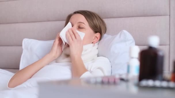 Een Europees uitziende vrouw met verkoudheid ligt op een bed met een verbandkeel. Behandeling van seizoensgriep en verkoudheid thuis. - Video