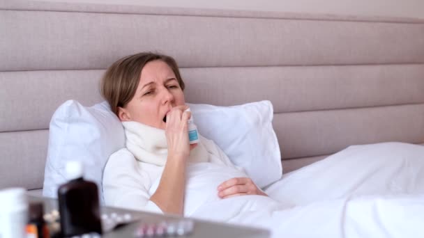 Een jonge vrouw met keelpijn ligt op een mooi bed en spuit haar keel. Behandeling van virus, verkoudheid en griep. - Video