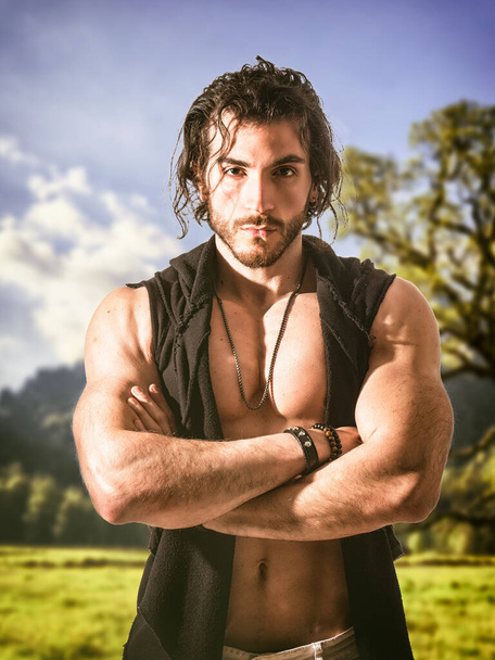 Confident, attractive muscular young man with open vest on muscular torso, ripped abs and pecs, outside in nature, with arms crossed on chest - Photo, Image