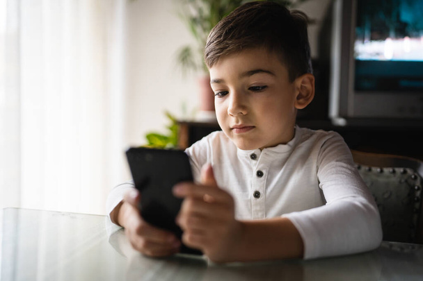un niño caucásico niño preescolar celebrar teléfono inteligente teléfono móvil en casa jugar videojuegos infancia y crecer concepto de adicción a la tecnología uso de la aplicación de teléfono inteligente para la navegación en línea o ver vídeo - Foto, imagen