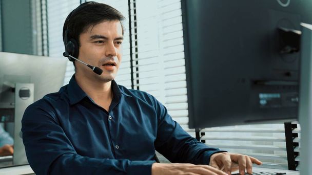 Young competent asian male call center agent working at his computer while simultaneously speaking with customers. Concept of an operator, customer service agent working in the office with headset. - Photo, Image