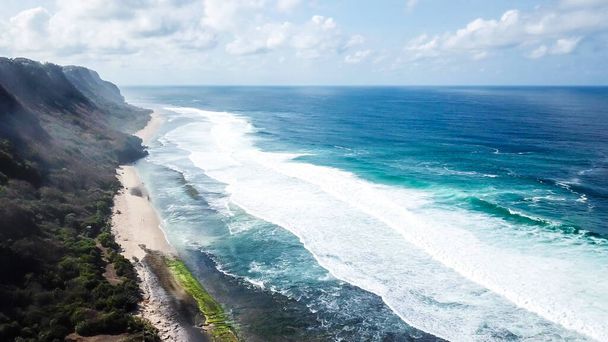 A done shot of Nyang Nyang Beach, Bali, Indonesia. The waves are rushing to the shore, making the water bubbly. The beach is covered with green algae, further on it's sandy. Tall cliffs on the side - 写真・画像