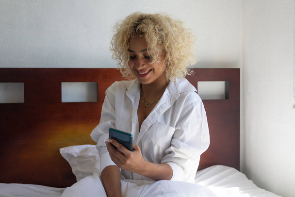 young hispanic girl using cell phone in bed smiling - Foto, Imagem