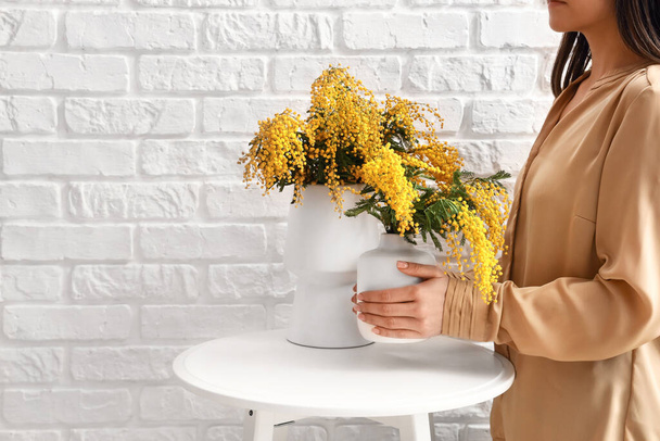 Woman holding vase with mimosa flowers on table near light brick wall in room - Foto, Imagen