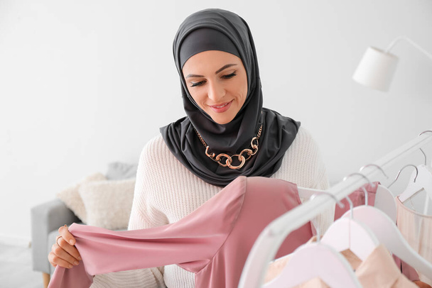 Beautiful Muslim woman near rack with clothes at home - Fotó, kép