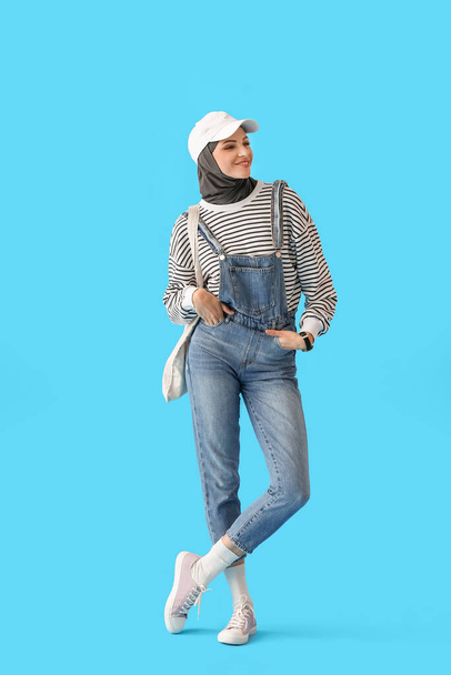 Stylish Muslim woman in cap with bag on blue background - Foto, afbeelding